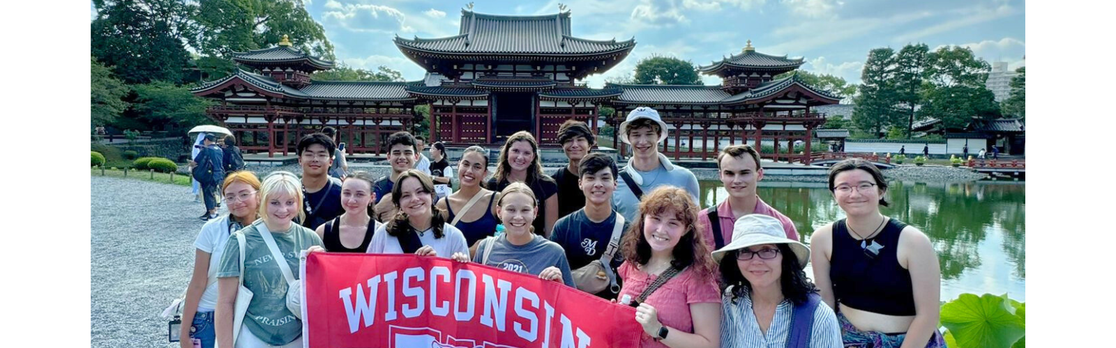 A group of students holds a Wisconsin flag and stands in front of a body of water, with a building with pointed roofs on the other side. 